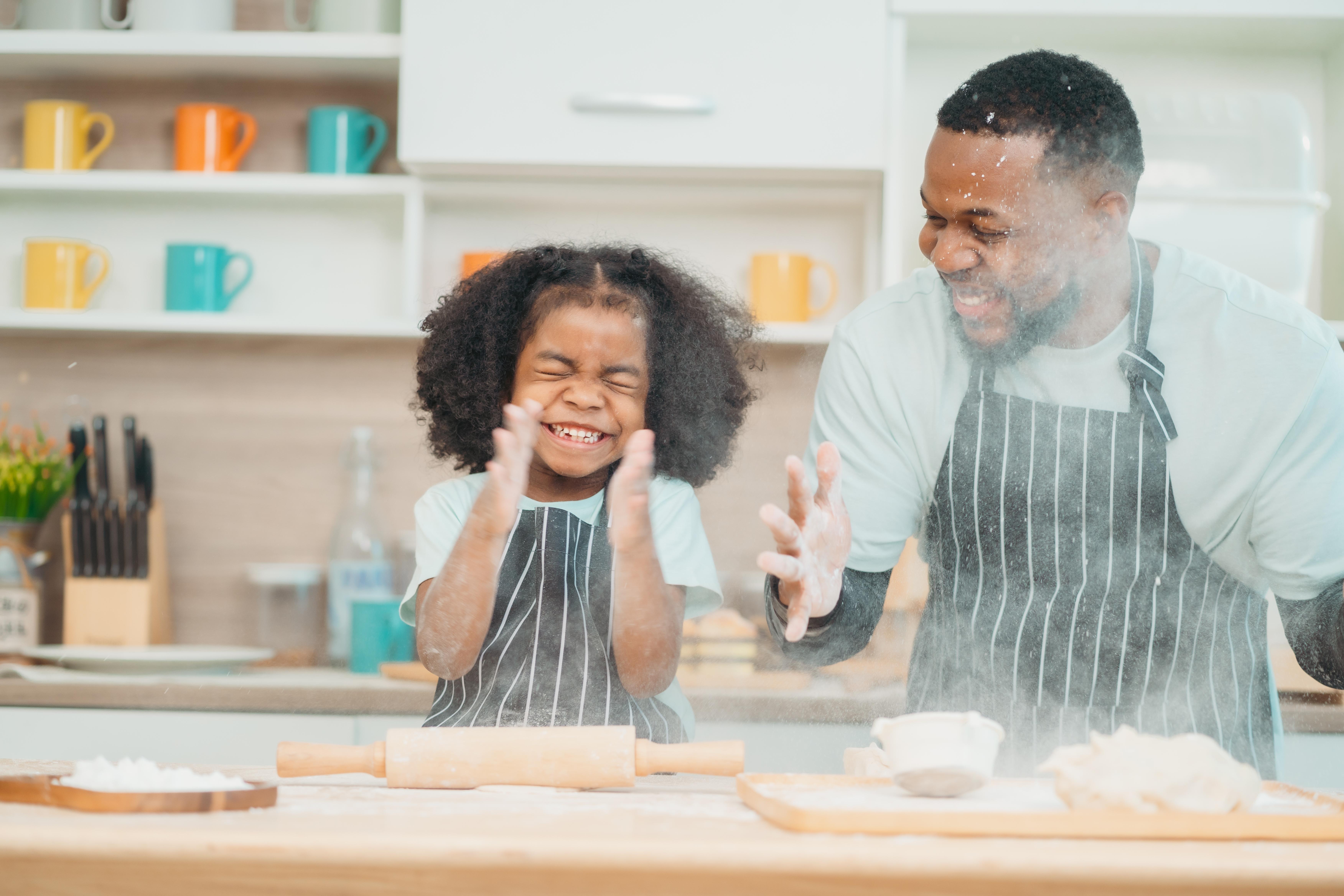 A father and young daughter wearing matching aprons bake together in a bright kitchen, both smiling as they work with flour and a rolling pin.