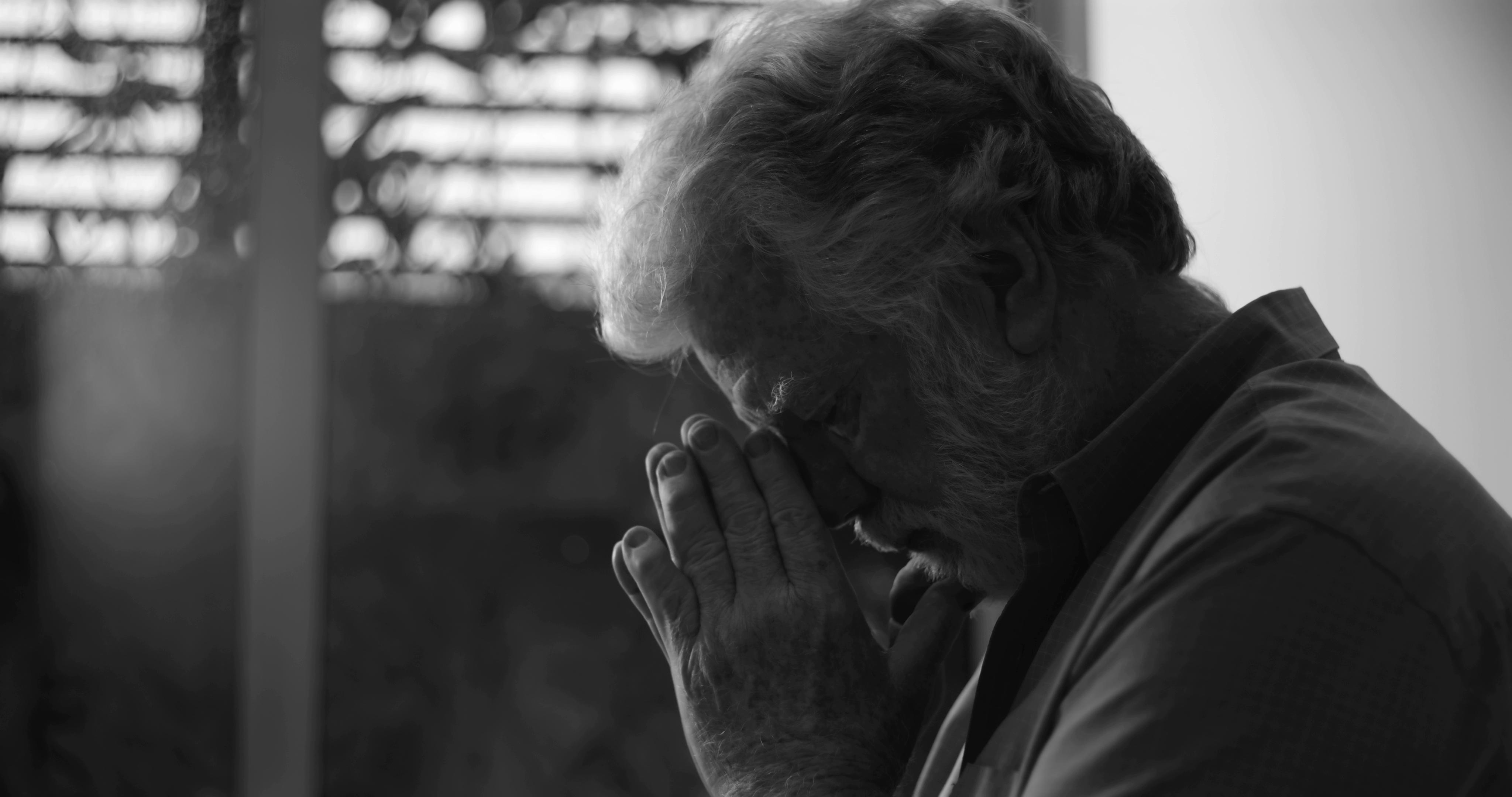 An elderly man with gray hair holds his face in his hands in a contemplative pose, photographed in black and white.