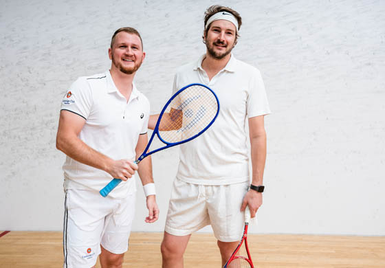 Two guys on the squash court, holding their racquets and smiling at the camera