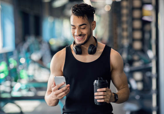 Fot, young man using his phone on the gym floor, with his protein shake in one hand, phone in the other, and headphones around his neck