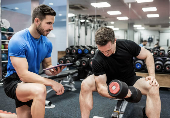 Young male trainer, kneeling next to a male client who is doing bicep curls. Trainer is holding a tablet. 